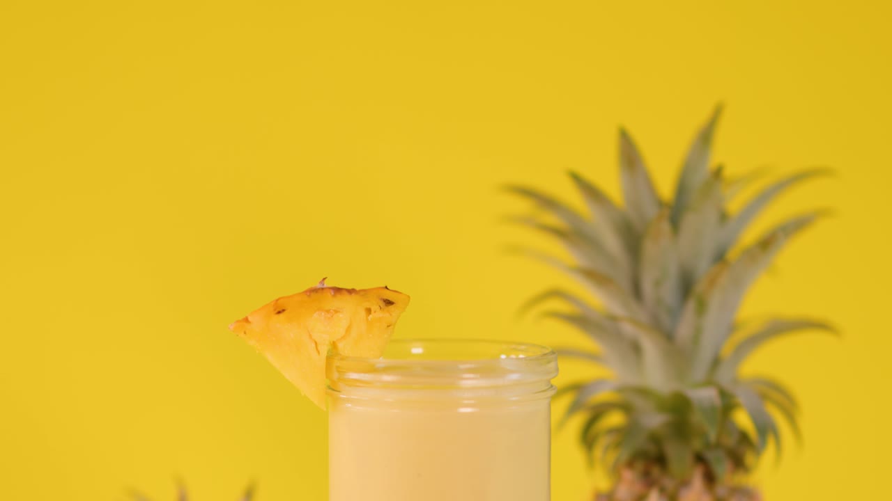 Hand places pineapple-shaped decoration on smoothie glass, bright yellow background, vibrant tropical atmosphere