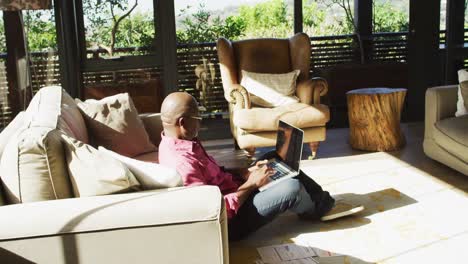 African-american-senior-man-sitting-on-living-room-floor-in-the-sun-working-using-laptop