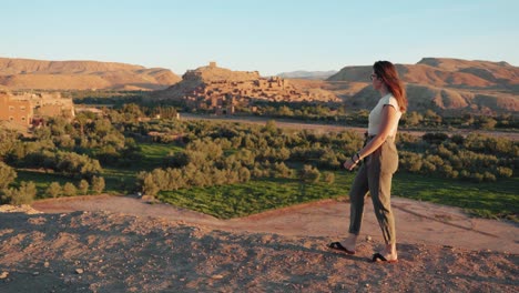 Wide-view-of-young-caucasian-woman-walking-and-looking-around-through-desert-landscape-at-sunrise-in-Ait-Ben-Haddou,-Morocco