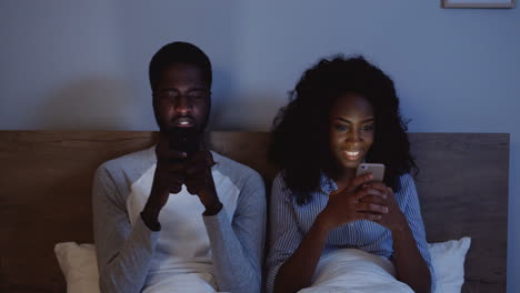 American-Couple-Laughing-While-Looking-At-Their-Cell-Phones-In-Bed-Before-Bedtime