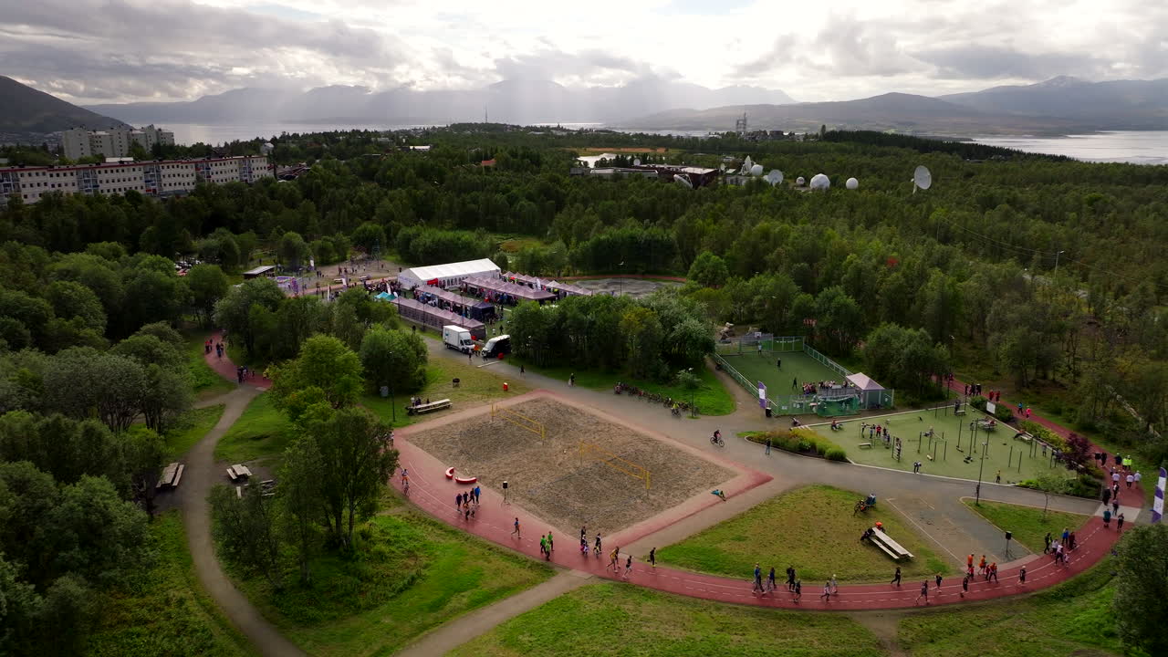 Aerial view of cancer Life race charity relay event with crowds walking and running around a track in Tromsø, Norway, surrounded by trees, sports facilities and dramatic Arctic scenery