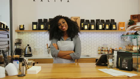 young african american female barista posing for the camera and crossing arms behind the bar of a coffee shop