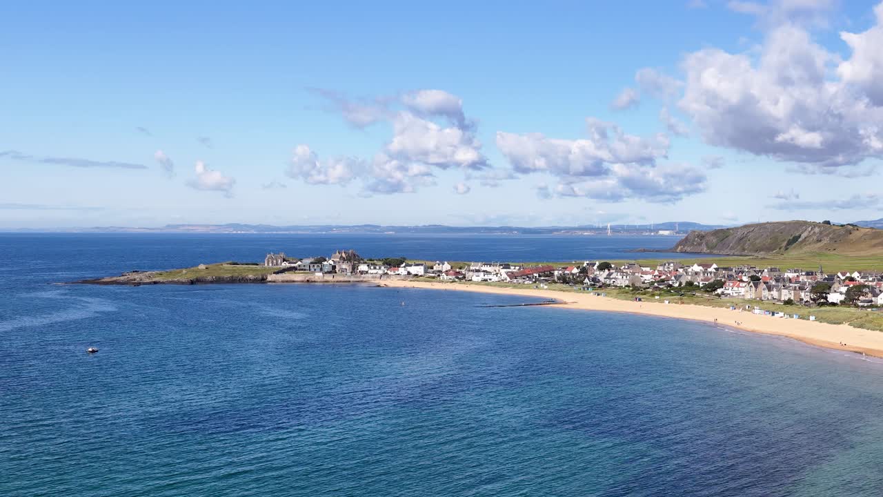 Drone glides toward Scottish coastal village, revealing blue sea, sandy shoreline, and clear sky
