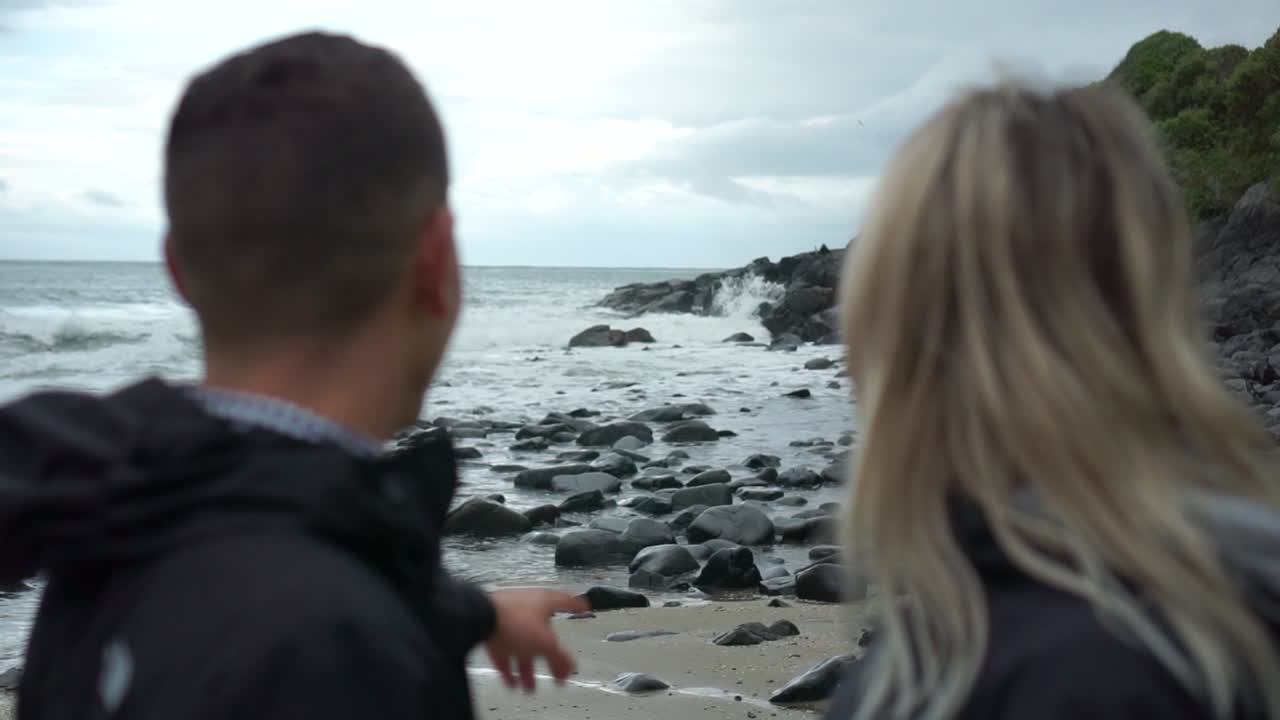 SLOWMO - Young couple on date - male pointing at ocean with his girlfriend on beach in New Zealand
