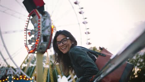 young stylish woman in glasses smiling cheerfully in front of a ferris wheel in amusement park