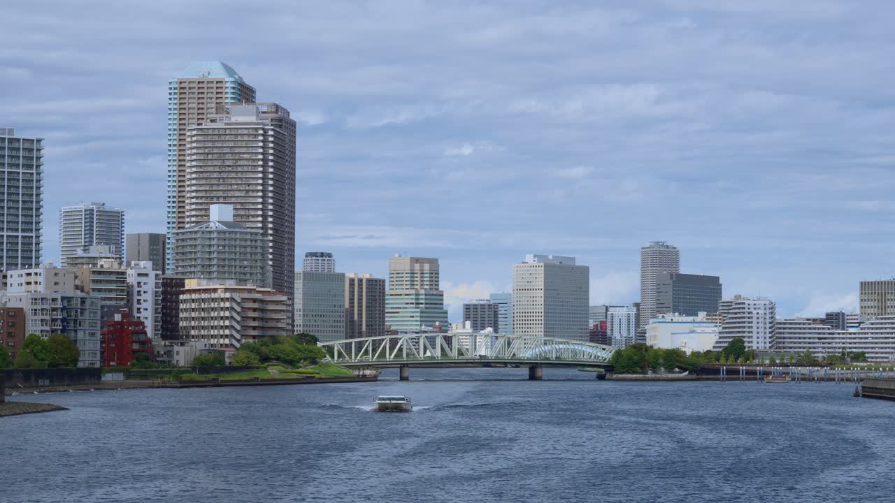 A peaceful shot of a passenger boat traveling on a river with the Tokyo cityscape and a bridge in the background