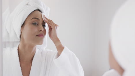 Biracial-woman-applying-cleansing-face-using-cotton-pad-looking-in-mirror-in-bathroom,-slow-motion