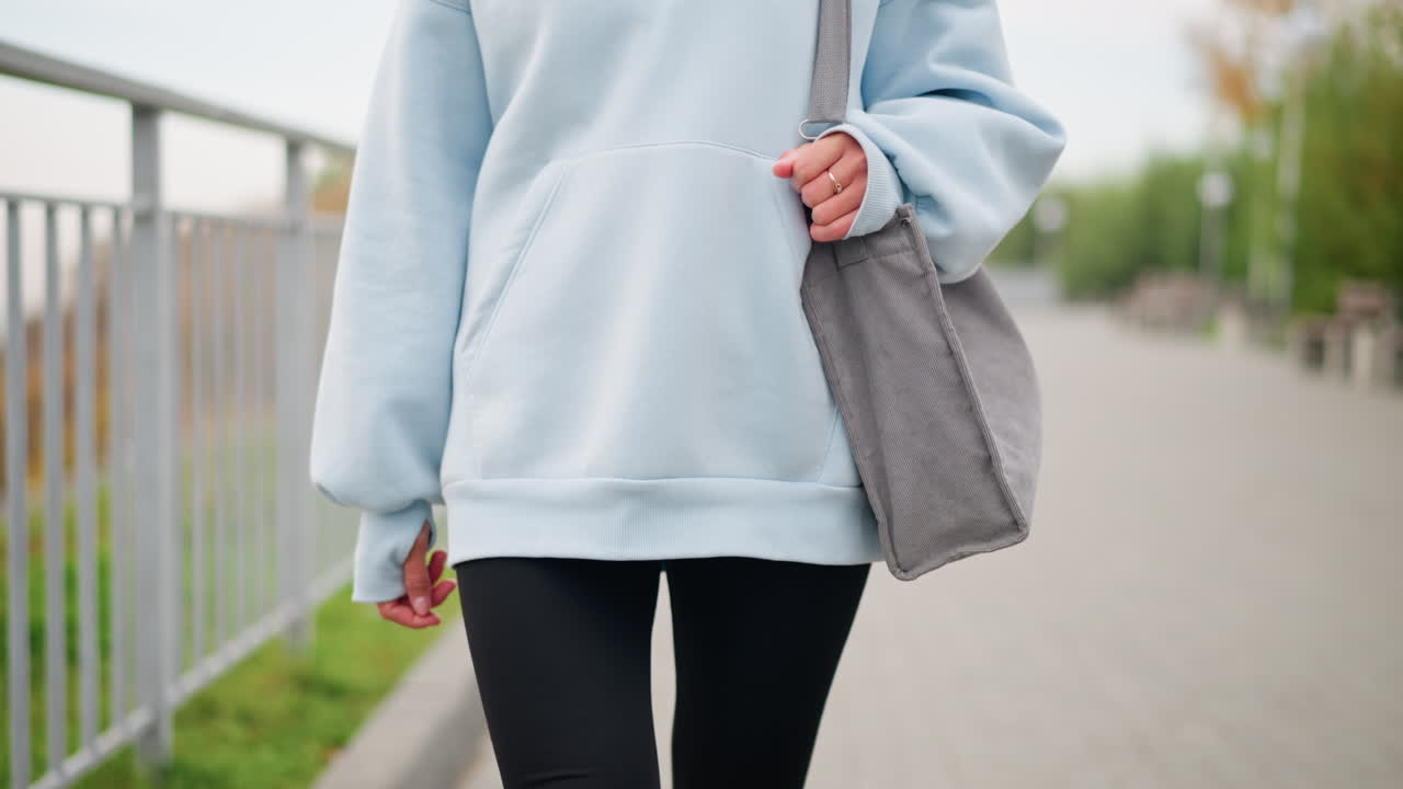 Close-up of young woman in casual attire walking with her bag and golden ring in hand, showcasing her relaxed and confident style, background features a blurred walking path and iron fence