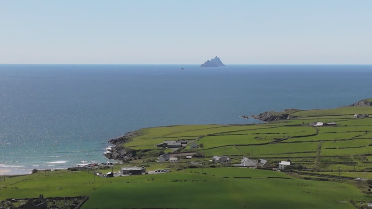 Static aerial view of distant Skellig Islands off the Irish coast. Lush green fields and calm ocean under a clear sky. Serene and expansive coastal landscape.