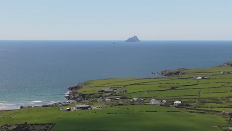 Static aerial view of distant Skellig Islands off the Irish coast. Lush green fields and calm ocean under a clear sky. Serene and expansive coastal landscape.