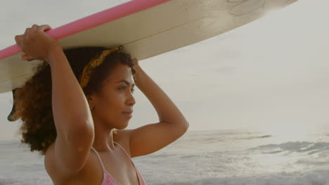 vista lateral de una mujer surfista afroamericana llevando una tabla de surf en su cabeza en la playa 4k