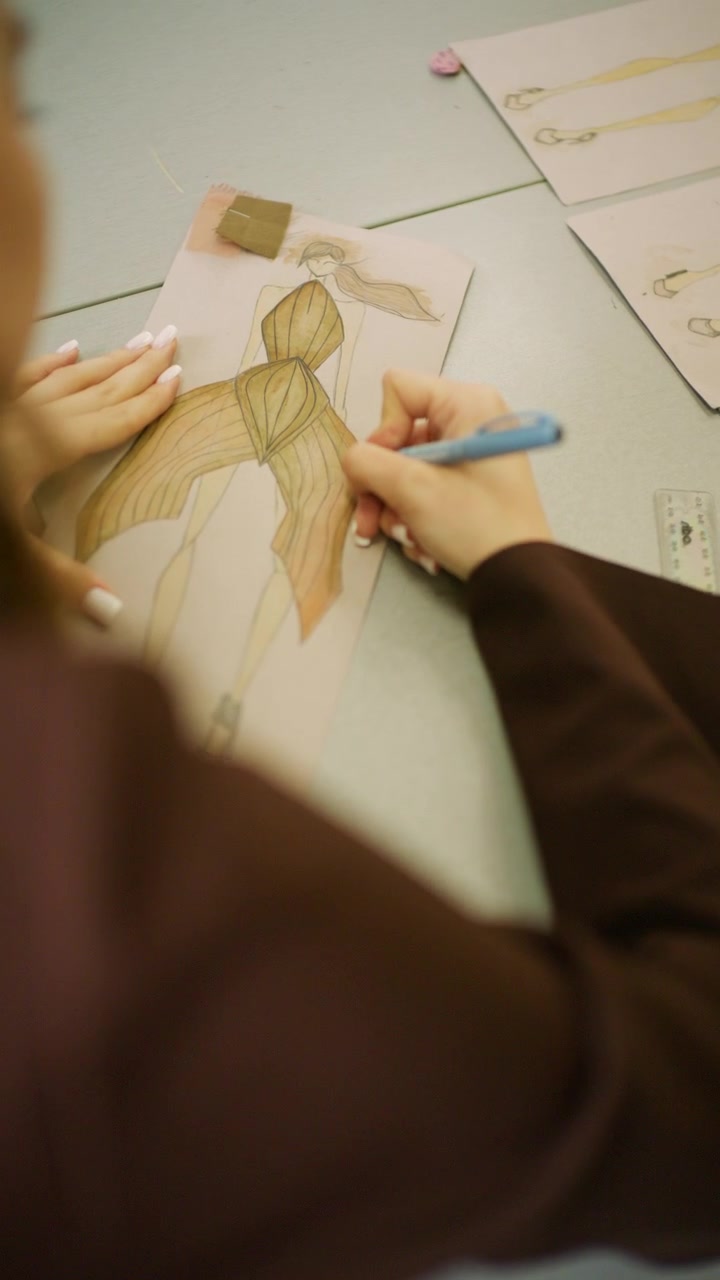 Close-up de um designer de moda desenhando uma ilustração de vestido estruturado dourado com lápis de cor em uma mesa de trabalho de estúdio.