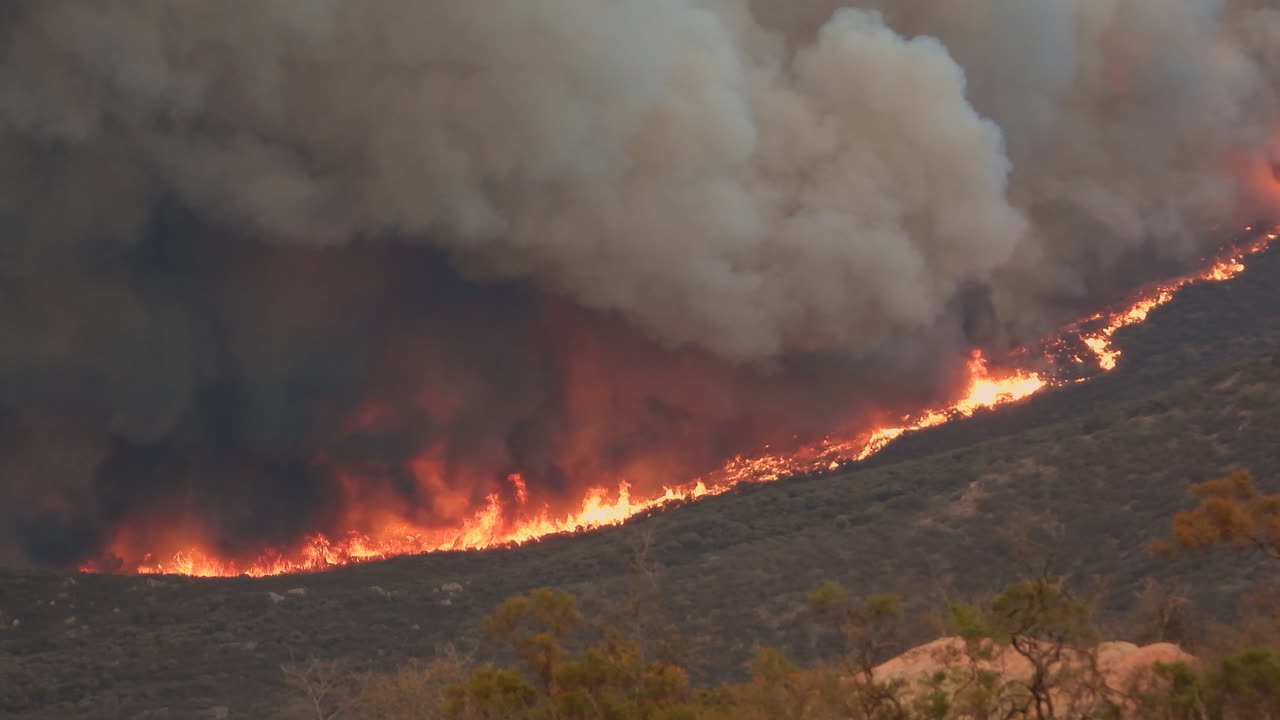 Incendio boschivo che brucia lungo un versante di montagna, con fumo nero e grigio che si innalza.