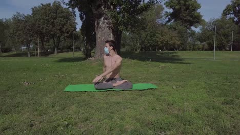 Hermoso-Joven-En-Forma-Con-Mascarilla-Practica-Yoga-En-El-Parque,-Hombre-Moreno-Haciendo-Ejercicio-Y-Pasando-Tiempo-Activo