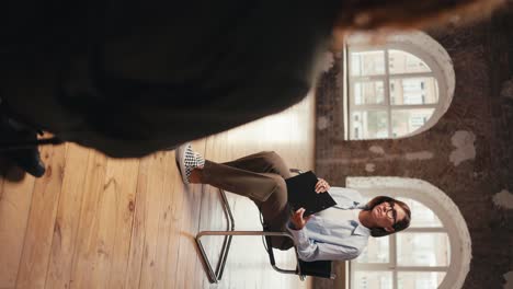 vertical video: a girl psychologist with a bob hairstyle and glasses in a blue shirt communicates with a participant in group therapy and interviews him