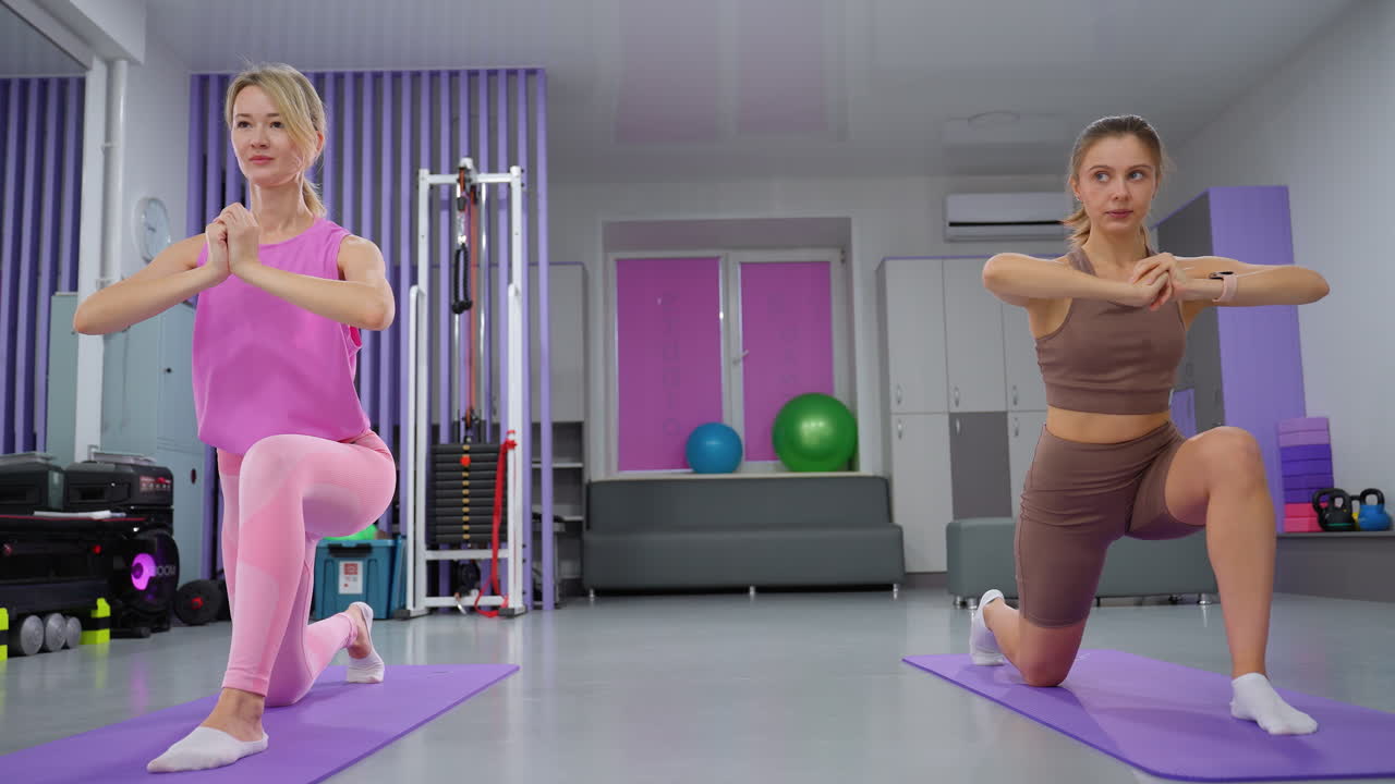 instructora femenina en marrón guía a la mujer en rosa a través del ejercicio de estiramiento de las piernas en el gimnasio, ambos se toman de la mano mientras se mueven arriba y abajo en esteras de yoga, centrándose en la fuerza y el equilibrio