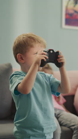 cute blond boy feels happy to annoy sister while playing video game with console. happy toddler jumps while preschooler girl sits on sofa closeup