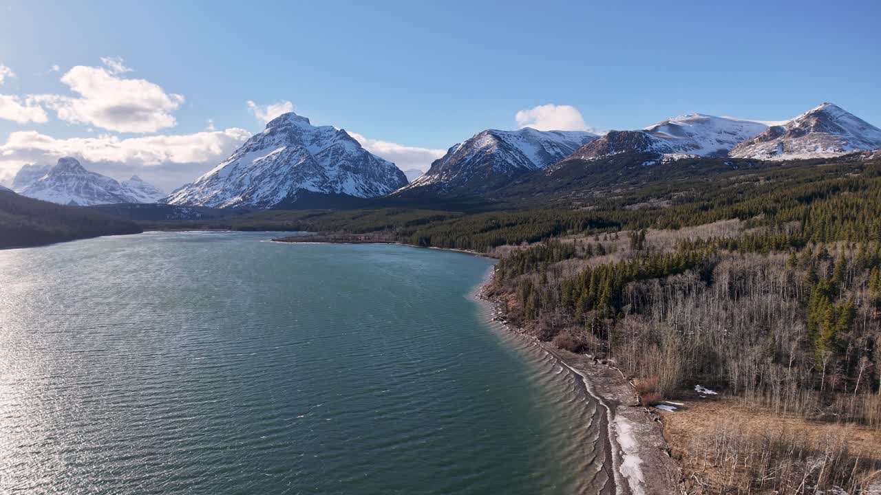 Riprese con drone mostrano un ampio lago di montagna dalle acque color smeraldo, delimitato da una riva boscosa e imponenti cime innevate sotto un cielo azzurro vicino a Whitefish, Montana.