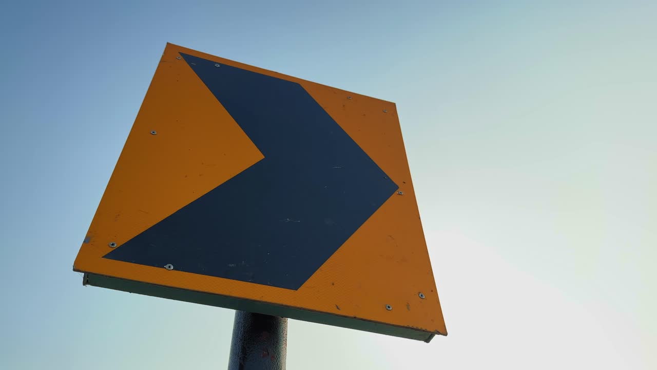 A close-up circling shot of a yellow highway turning marker with a black arrow, captured against a clear blue sky, symbolizing road direction and safety