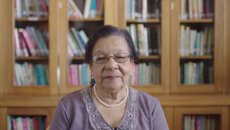 Retrato-De-Una-Elegante-Mujer-Anciana-De-Raza-Mixta-Sonriendo-Contenta-A-La-Cámara-En-El-Fondo-De-La-Biblioteca-Usando-Un-Collar-De-Perlas