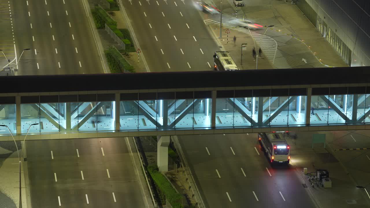 Dynamic nighttime timelapse capturing flowing vehicular traffic under a modern, illuminated glass pedestrian bridge. The glowing lights and blurred streaks create a vibrant urban scene