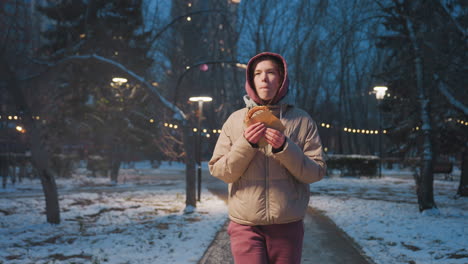young woman walking in outdoor park during winter, eating donut wrapped in napkin, enjoying snack while walking along snowy path with street lights and trees in background