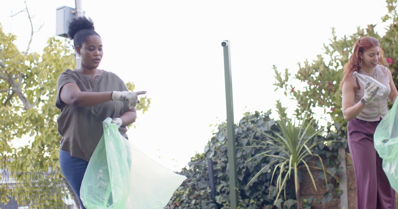 Two women spotting litter, wearing white gloves, picking bottles into green bag while cleaning yard