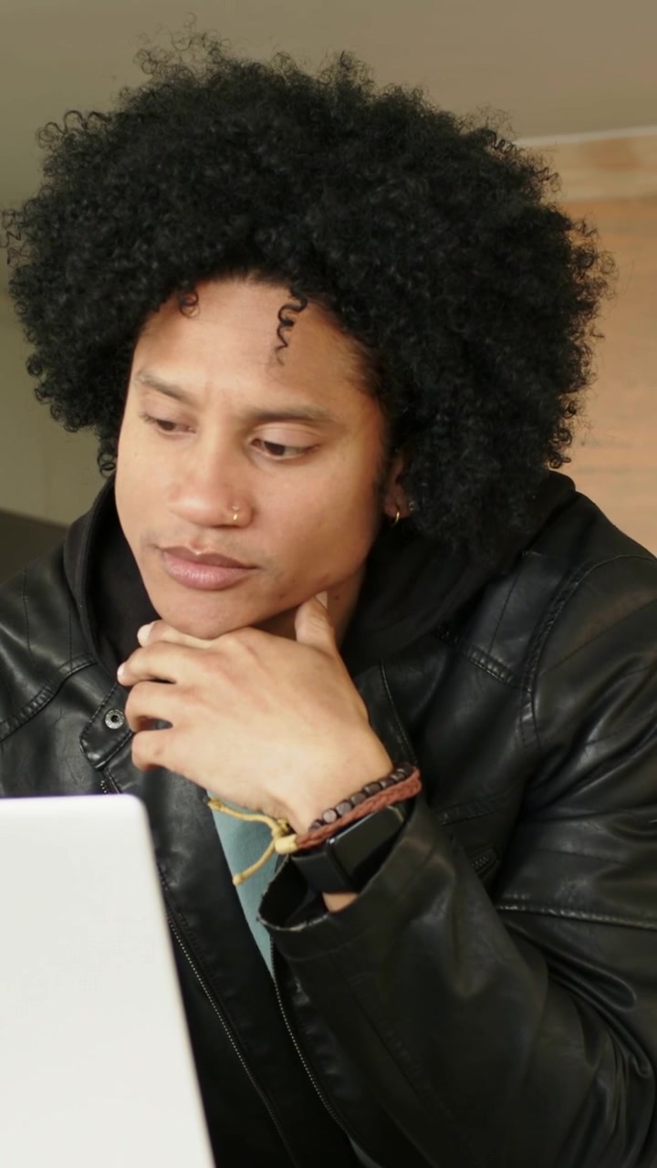 Vertikal video: Laptop i fokus, mann i skinnjakke og smartklokke lener seg og studerer hjemme.