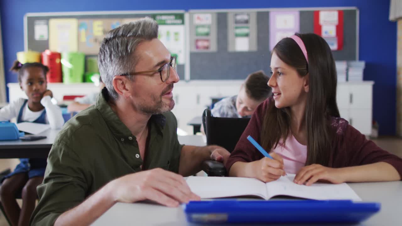 un maestro masculino diverso ayudando a una escolar sentada en silla de ruedas en el aula