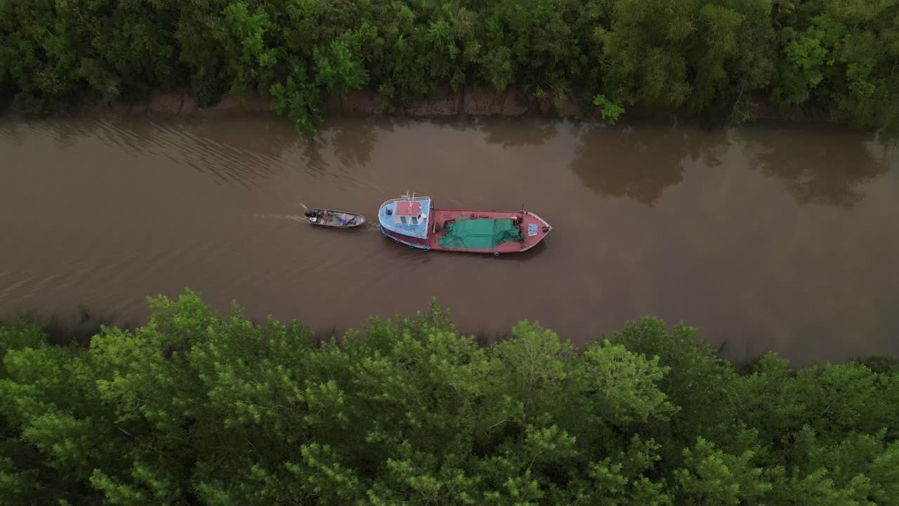 Cinematic drone side shot of ship carrying small boat on amazon river surrounded by green rainforest trees during sunrise