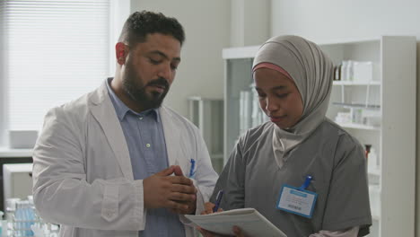 scientist talking to female assistant in hijab taking notes in laboratory