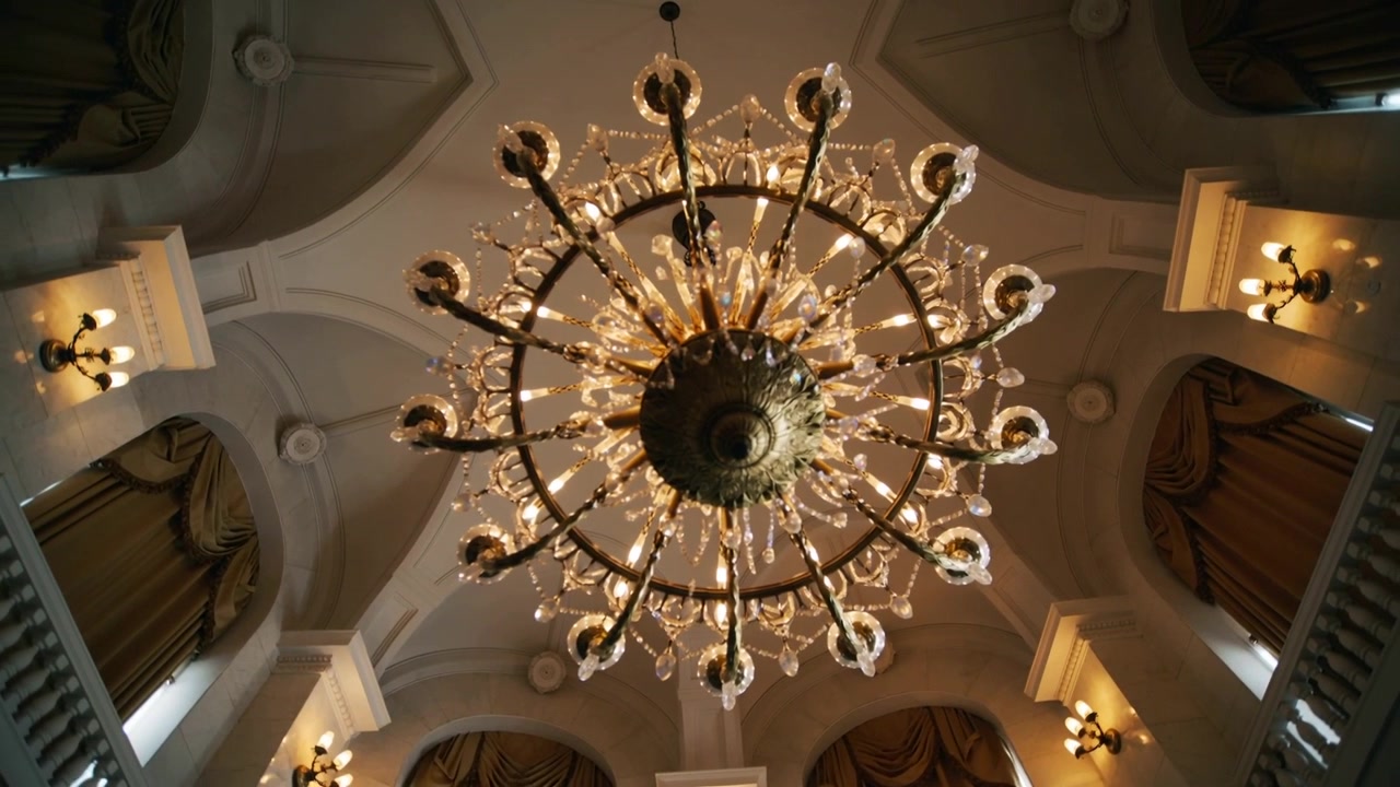La telecamera si inclina catturando un grande lampadario di cristallo con lampadine accese e modanature in gesso in una sala da ballo.