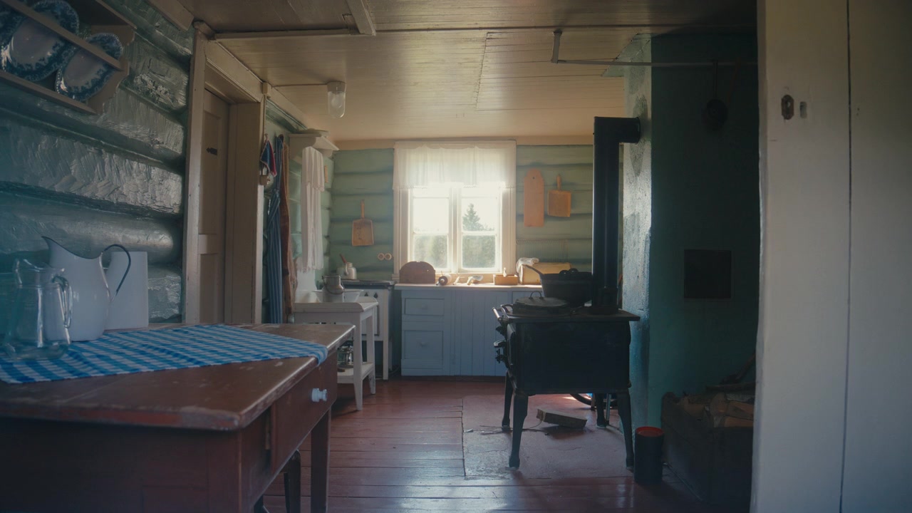La luce soffusa della finestra illumina una cucina rustica di baita con pareti di tronchi dipinti, mobili in legno e una stufa in ghisa, creando una scena interna calda e nostalgica.