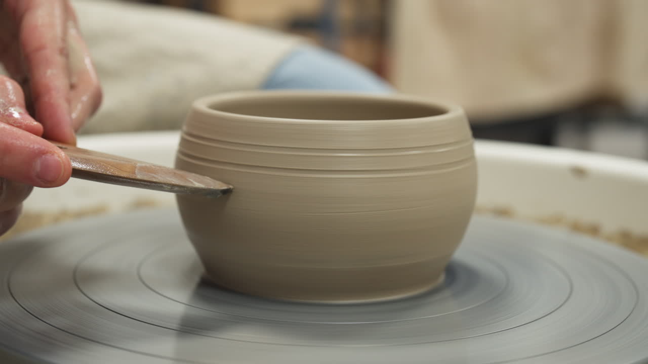 Medium shot of white hands making mug from clay with a piece of wood, interior