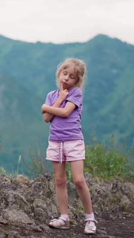 niño pequeño en traje de lila toca la mejilla con el dedo en el suelo de la colina de piedra fantaseando contra las montañas forestales en el eco resort en un día nublado en cámara lenta