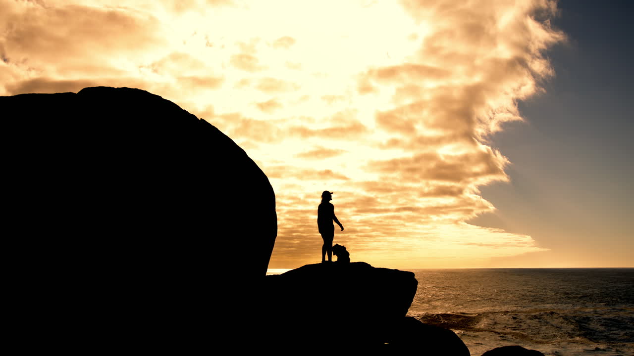Silhouette of a person and a dog on rocks at sunset