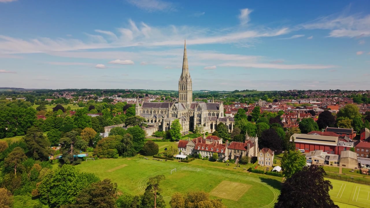 Ripresa aerea con drone della Cattedrale di Salisbury nel Wiltshire, Inghilterra, che mostra la cattedrale gotica medievale e la famosa guglia circondata da prati verdi, alberi, edifici storici e lo skyline più ampio della città