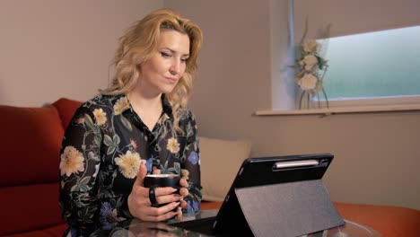 mujer de pelo rubio viendo su tableta sosteniendo una bebida caliente