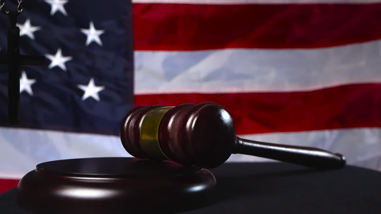Brown wooden ceremonial gavel hammer hitten on wooden podium by a judge justice hand while a holy cross of faith moves in the foreground and American United States flag is in the background