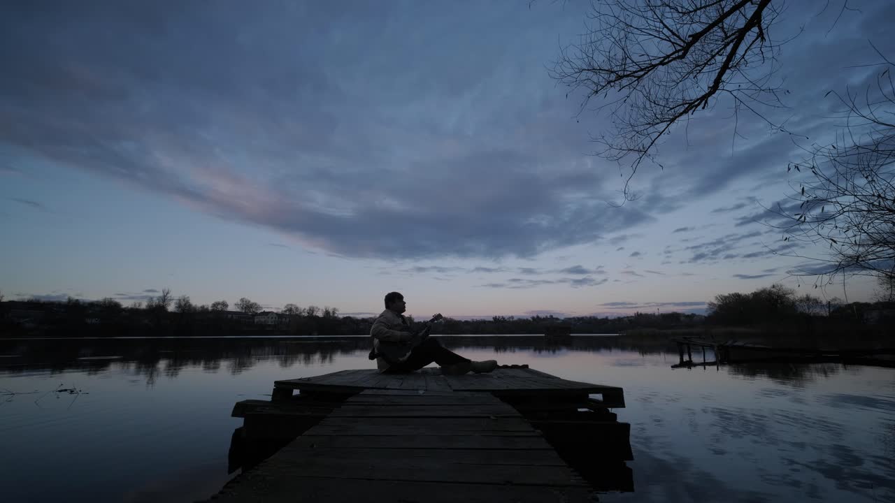 Silhouette of musician in black playing guitar sitting on pier embankment on sunset
