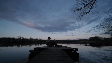 silhouette of musician in black playing guitar sitting on pier embankment on sunset