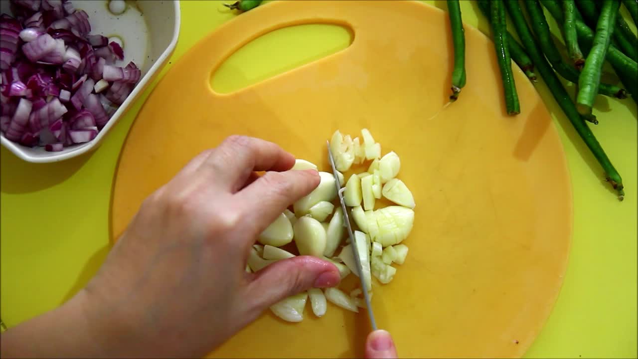 Overhead or flatlay steady video shot of a person chopping garlic with a knife on a chopping board