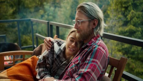 A-girl-hugs-a-blond-guy-with-glasses.-A-couple-is-resting-on-a-devan-wrapped-in-a-blanket-on-the-balcony-of-a-country-house-overlooking-the-mountains-and-forest