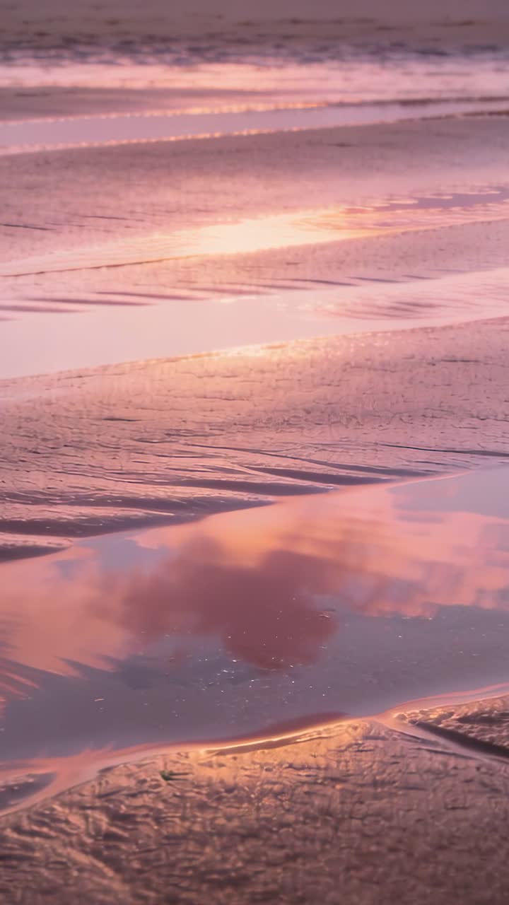 Vertical video: Rippling tidal pool reflecting pink clouds and sparkles on wet sand during sunset