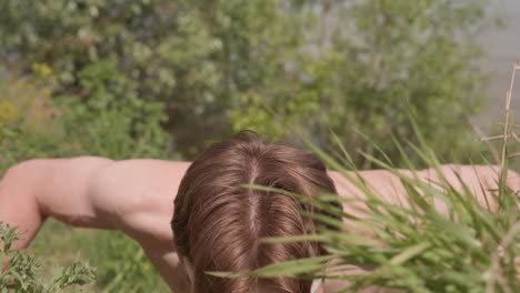 front view of shirtless soldier placing hands firmly on riverside grasses under sunlight with tactical gloves and camouflage face paint as he prepares to climb muddy bank near flowing water