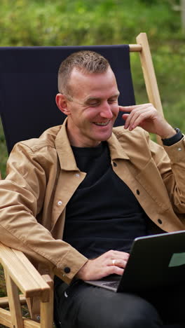 caucasian man in brown shirt sitting with laptop on his laps in the garden. relaxed freelance worker talks to someone laughing cheerfully. vertical video