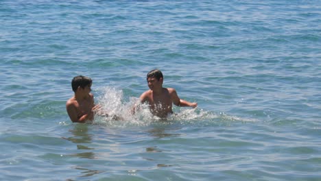 Caucasian-10-and-8-years-old-brothers,-teasing-each-other-with-splashing-water-at-seashore,-medium-100fps-slow-motion-footage