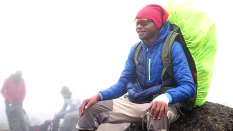 a black man sits and smiles with a backpack and jacket on a boulder during heavy fog