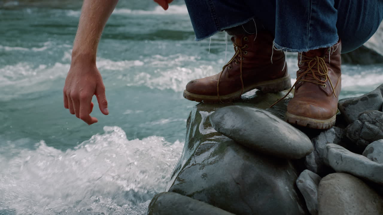 Man zit op rots bij beek. Mannelijke hand raakt fris water uit rivier aan