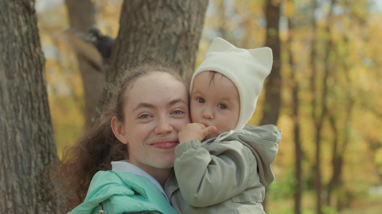 Ragazza in un parco autunnale che tiene in braccio un bambino, tenero momento familiare tra giovane donna e neonato vicino a un albero, giacca calda e berretto, coccole e sorrisi, sfondo di foglie dorate, ritratto spontaneo che trasmette affetto